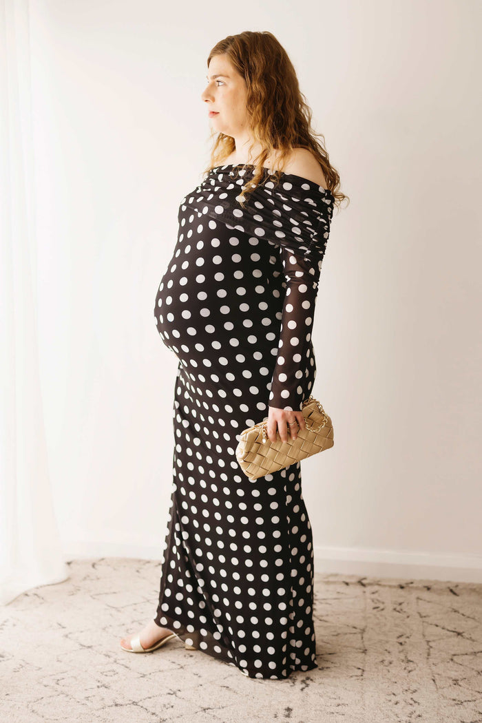 A pregnant lady wearing the Mama Rentals AFRM Thelma Off Shoulder Mesh Maxi Dress - Black in a studio.