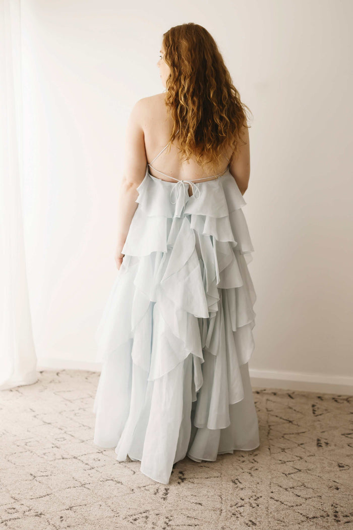 A pregnant lady showing the back design of the Mama Rentals Aje Pandorea Layered Maxi Dress - Silver Blue in a studio.