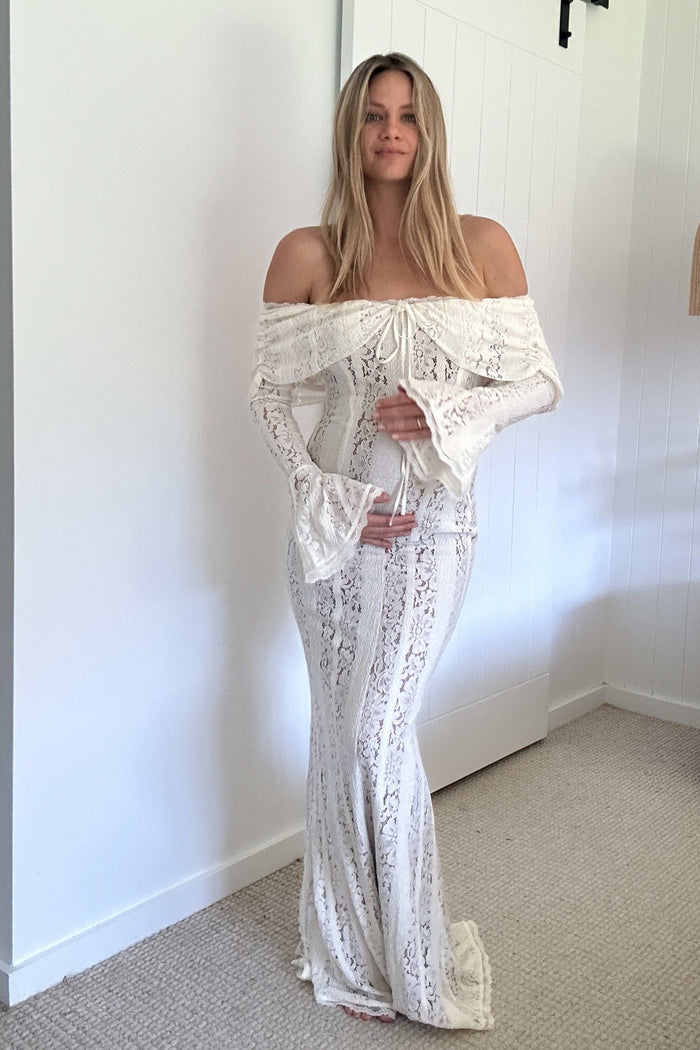 Woman wearing the Mama Rentals House of CB Belle Vintage Cream Floral Lace Off Shoulder Maxi Dress in a room with a plain white wall.