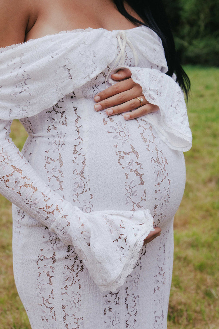 Pregnant woman embracing her belly in Mama Rentals’ House of CB Belle Vintage Cream Floral Lace Off Shoulder Maxi Dress, surrounded by soft greenery and trees.